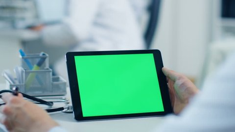 Close-up of a Male Doctor Uses Tablet Computer with Green Screen, He also Holds Pen and Makes Notes, His Assistant Works in the Background. Office is  Modern. Shot on RED Cinema Camera 4K (UHD).