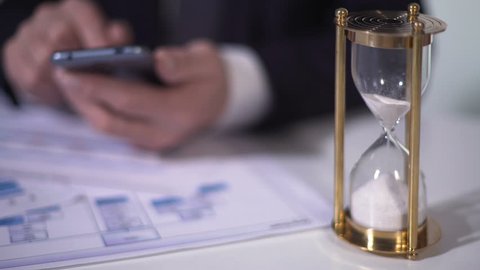 Time running out with sand in hourglass, defocused man using smartphone for work