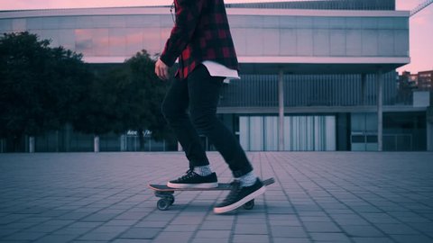Fashionable rider in hipster red plaid shirt and black jeans just skates on his longboard at twilight time with beautiful paster lightning, camera follows him