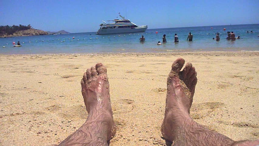 Man's feet in sand with yacht in tropical location. Chileno Bay in Cabo, Mexico.