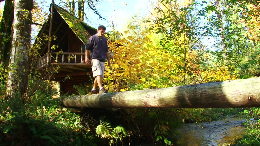 Log cabin set back in beautiful forest with river brook, man crosses wooden