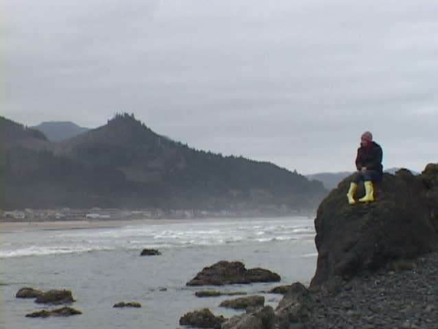 Woman at Haystack Rock in Cannon Beach, Oregon sits looking out by ocean. 