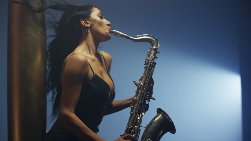 Young, beautiful, sexy, brunette woman dancing and playing golden saxophone on dark backg...