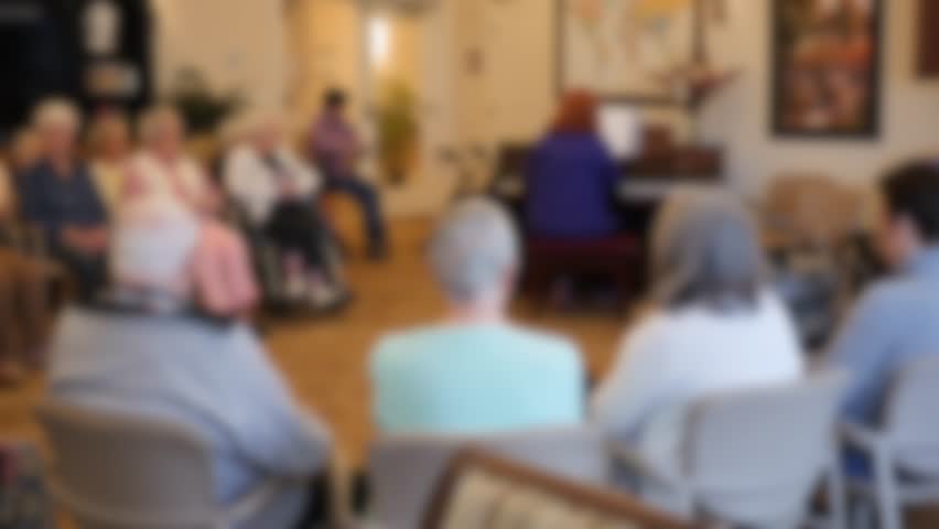 An out of focus shot of elderly people in an assisted living home listening to someone play the piano