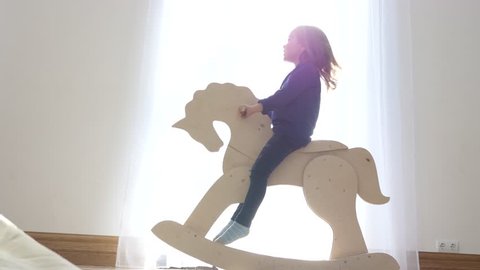 Little Child Girl Sway On A Wooden Rocking Horse For Children Near Bright Window