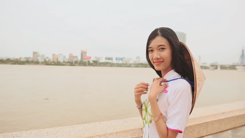 Vietnam girl in the national costume and dress Ao Dai posing on the waterfront of the city of Da Nang. On the back of the cap Non La.