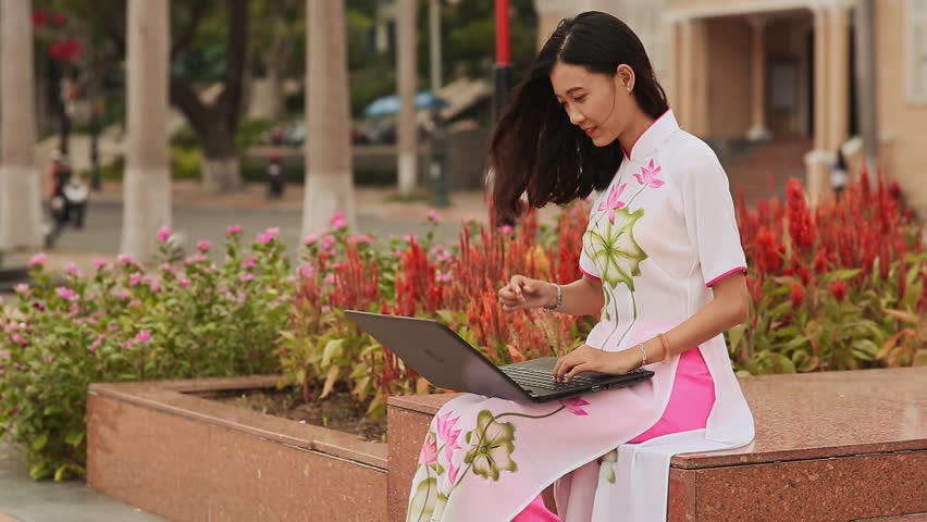 Pretty Asian girl sitting with a laptop in the street city in national dress Ao Dai