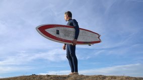 Surfer standing on sand dune with surfboard, looking at the sea
