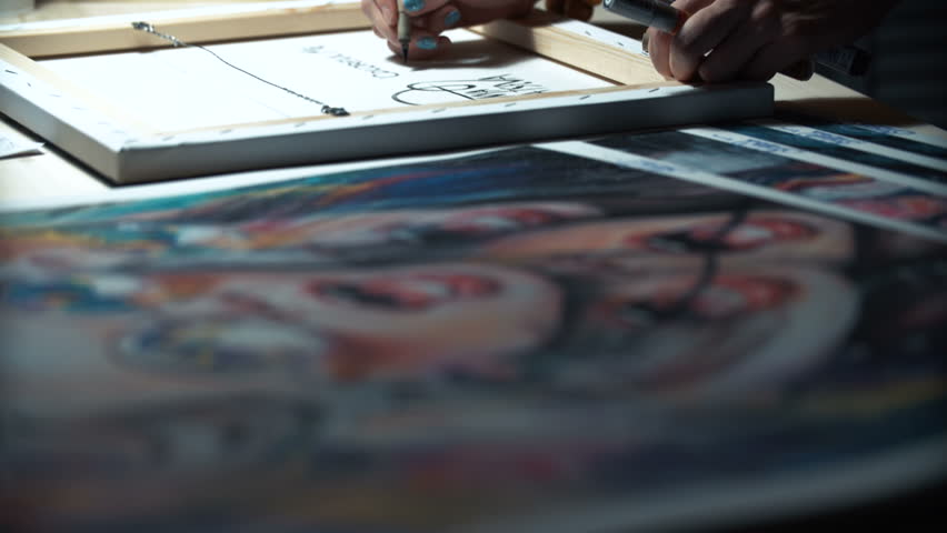 Girl artist signs some of his works. Painted sign of a master. Canvas on a stretcher signature black marker. On the desktop, next are other painted art prints. Close up