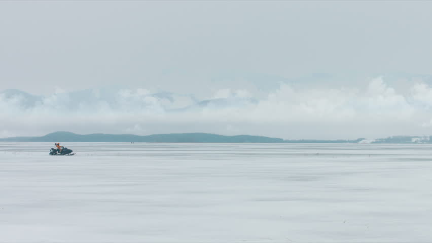 WIDE 2 persons driving sports snowmobile on a large frozen lake, mountains in the background. 4K UHD 60 FPS RAW edited footage