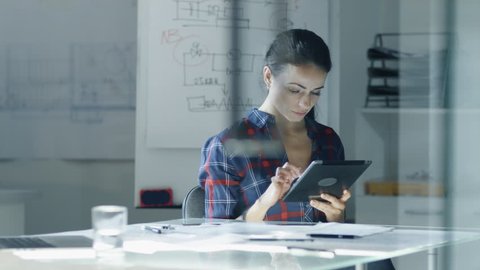 Female Design Engineer Sits at the Glass Table in Her Office, Works on a Tablet Computer, Blueprints Laying on Her Desk. In the Background Whiteboard with Diagrams and Sketches. 