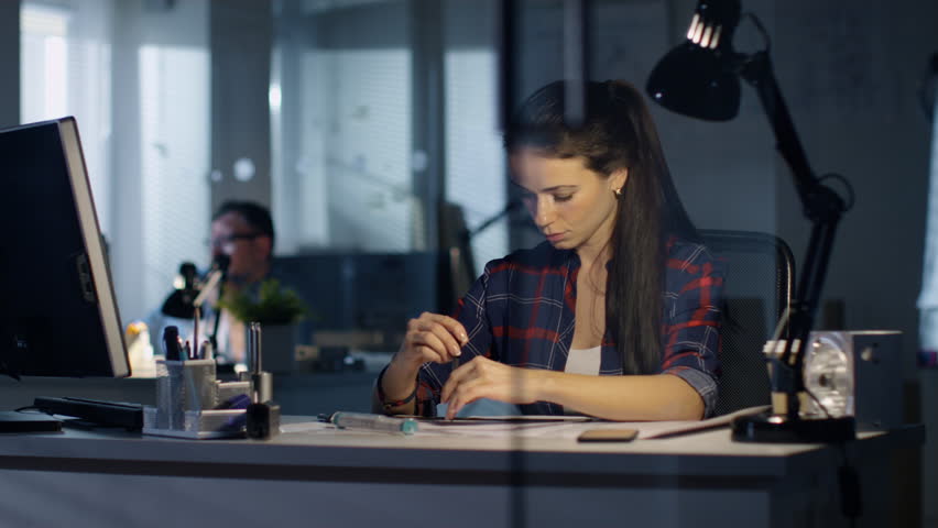 In Late Night Office Female Design Engineer Drafts with the Help of Rulers and development.  Colleagues Work in the Background.Shot on RED Cinema Camera 4K (UHD).