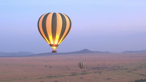 AERIAL: Safari hot air balloon flying above vast savanna plain rolling into the distance in Serengeti National Park at purple light dawn. Tourists traveling in air over African wilderness at sunrise