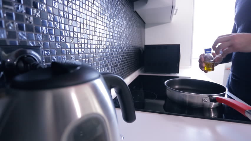 In the kitchen man pours oil onto the hot pan on an induction cooker. From a small bottle of Male slowly pours the oil into a container with a red handle for that day to cook dinner. In the stylish