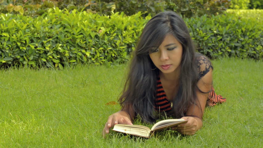 A beautiful young Asian woman reading a book, while relaxing, lying on the green