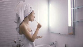 Beautiful young woman with towel wrapped around her head brushing teeth and dancing while standing against mirror in bathroom.