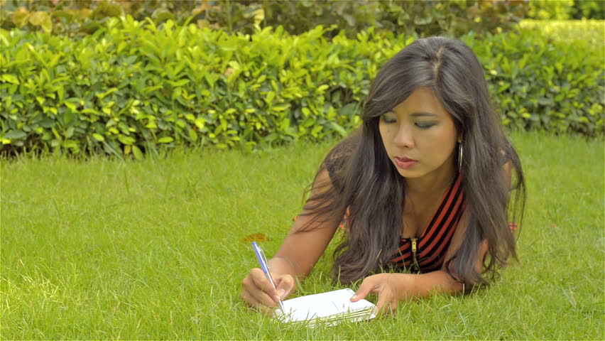 A beautiful young Asian woman,lying on the grass in a lush park, writing in her