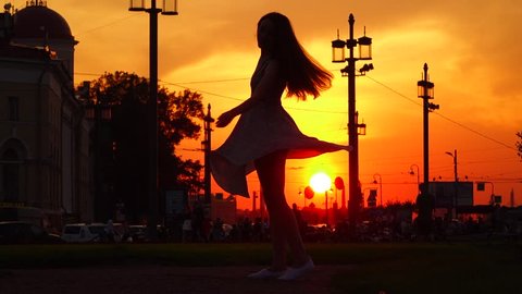 Merry young woman happily turn around in dance, short light dress skirt fly around, black silhouette against vivid orange sunset, urban place. Slow motion shot, live is good concept