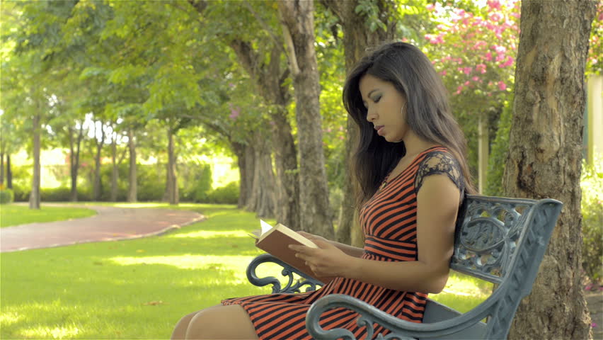 A beautiful young Asian woman reading a book, while relaxing, sitting on a park