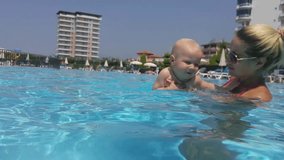 Mother dip baby into water of swimming pool near hotels close up at day, mobile phone video.