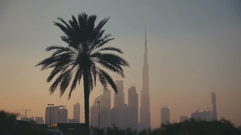 Sunset in Dubai: Palm silhouette of the Burj Khalifa and other skyscrapers, past the camera passing cars. Sunrise Sunset Tall Building Burj in Dubai. Skyline Cityscape City