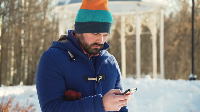 young man in blue coat and colorful hat with beard typing text in smart phone, look up and stare to camera without smile, with steam from the mouth because of cold winter day under the sunlight