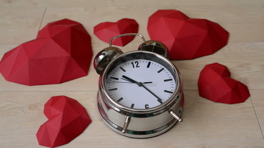 Big alarm clock with red polygonal paper heart shapes over wooden bachground, top view
