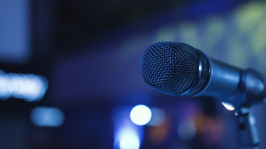 Microphone on the Stand is Standing on the Stage, Close-Up Microphone on the Background of the Auditorium, Spotlight, Backlight People Gather in the Hall and Sit on Seats, Waiting For Performances 4k Royalty-Free Stock Footage #23853730