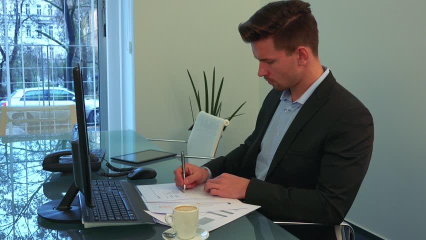 A young, handsome man writes on papers at a desk in an office, a window in the background