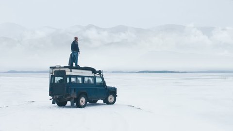 WIDE Caucasian male standing on the roof of old 4x4 off-road vehicle, spreading hands and enjoying the view of mountains over large lake. 4K UHD 60 FPS RAW edited footage