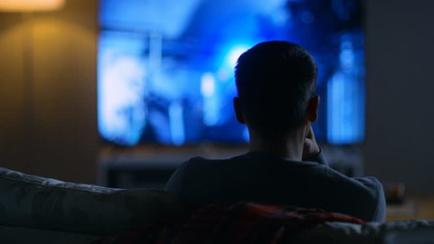 Back View of a Man Sitting on a Couch Watching Movie on His Big Flat Screen TV. Shot on RED EPIC-W 8K Helium Cinema Camera.