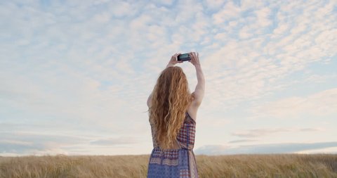 Rear view of attractive red head woman taking photograph wheat field using smartphone capturing epic nature background
