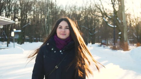 follow after young attractive woman with long dark hair in black coat and knitted scarf who look at camera, go around and smile while walking in the snowy park under the sunlight