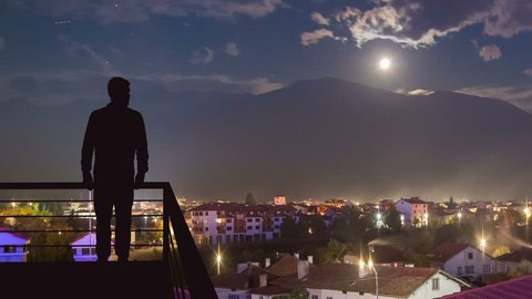 The man stand on the balcony on the background of the evening town. Time lapse 