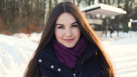 young attractive woman with long dark hair in black coat and knitted scarf look at camera and blow kisses while walking in the snowy park under the sunlight