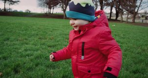 Happy kid in red jacket running and smiling in park on green grass. 4k video. Slow motion.