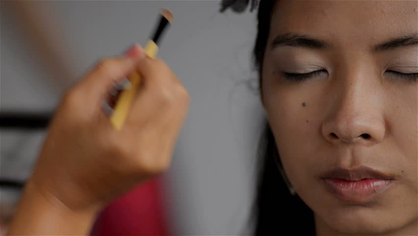 Makeup artist applying eyeshadow to the eyelids of an Asian woman.