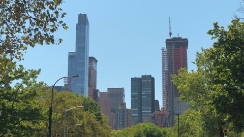 LOW ANGLE VIEW: Driving along the tree avenue on beautiful sunny summer day in New York City Central Park towards tall modern glassy skyscrapers in Midtown Manhattan. Luxury real estate in metropolis