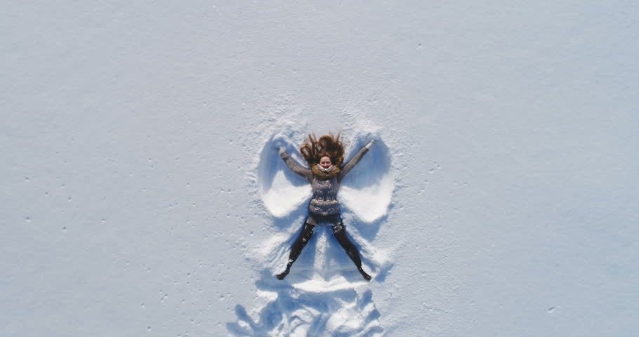 Young cheerful woman is lying on snow and is making snow angels. Slow motion. Aerial view with rotation.