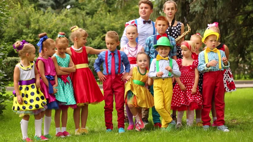 Fifteen children of different ages in bright clothes standing in park and waving