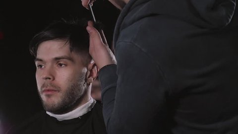 Making haircut look perfect. Young bearded man getting haircut by hairdresser while sitting in chair at barbershop, hairdresser styling.
