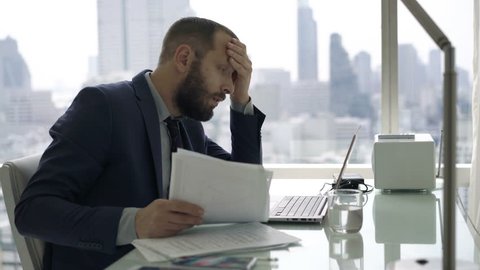 Sad, unhappy businessman working with documents and laptop by desk in office
