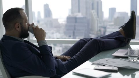Young businessman relaxing, drinking water and admire view sitting by desk in office
