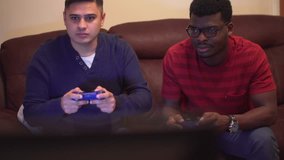 Two gamers, males competing on a video game console on sofa. Young adult asian male and black male. Competitive game between friends, one celebrates when he wins. Evening at home on sofa, casual dress
