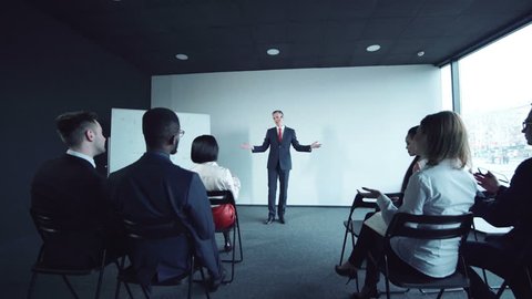 Group of young multiethnic businesspeople listening presentation in a meeting from adult man in a suit, side view. At the end of speech all people standing and start to a applaud