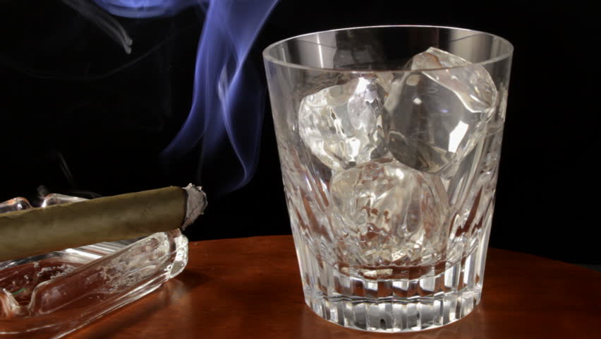 Cigar smoke rises from ash tray next to glass of scotch