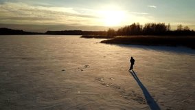 Aerial view: One man is skating on the ice of frozen river during beautiful sunset. Drone is following him and making the video. Man is enjoying the sunny day. 