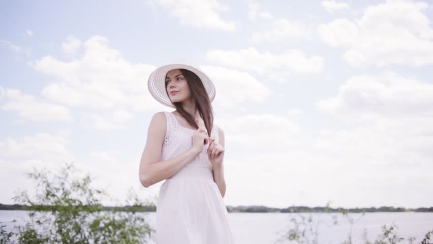 Young pretty brunette woman with hat near water with sky full of clouds. Shallow depth of field. Holding her hair with hands