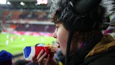 Teenager in a fur hat eating a hot dog at the stadium during a football match