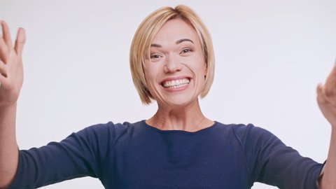 Blonde beautiful middle-aged woman loudly saying wow showing ok and smiling at camera standing on white background in blue sweatshirt